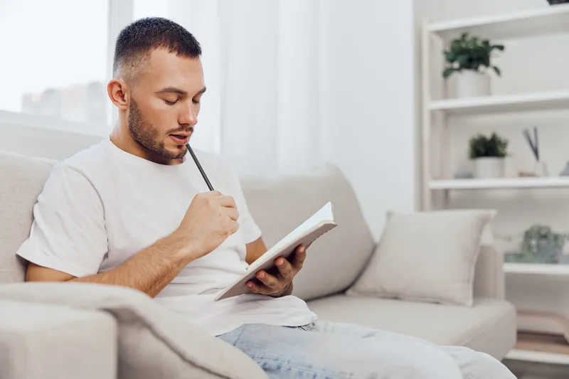 Focused man writing in a notebook while sitting on a cozy sofa, surrounded by bright indoor plants, conveying creativity and inspiration in a modern home setting.