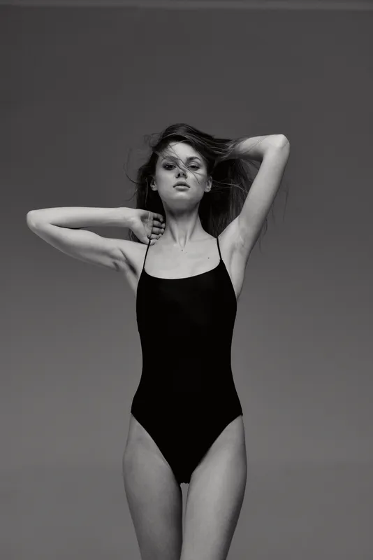 Black and white portrait of a young woman in a black swimsuit posing with her hands in her hair against a plain background.
