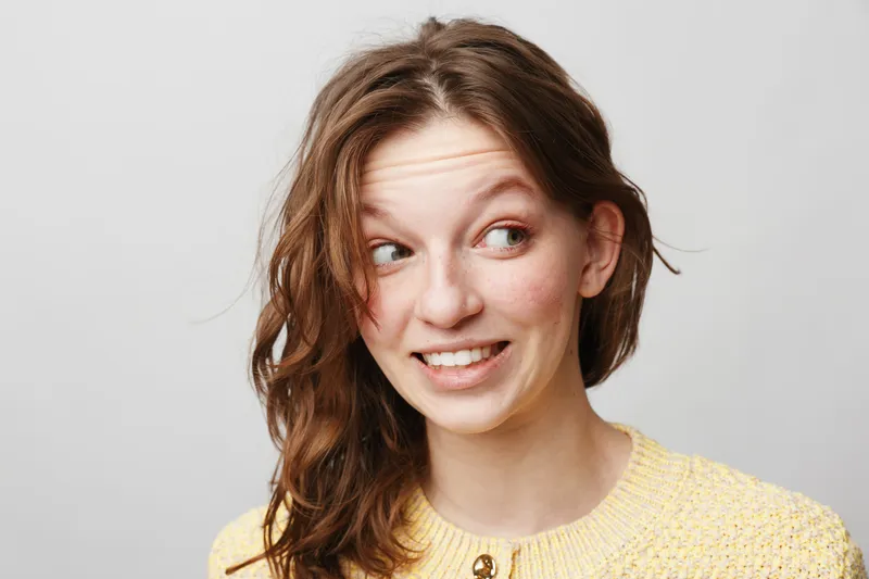 Young woman with wavy brown hair wearing yellow sweater looks sideways with a smile on light grey background. Concept of casual style, natural beauty, and positive emotions.