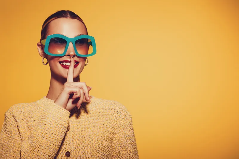 Stylish woman in large blue sunglasses and yellow sweater makes silence gesture with finger on lips. Bright yellow background creates vibrant and playful mood for fashion concept.