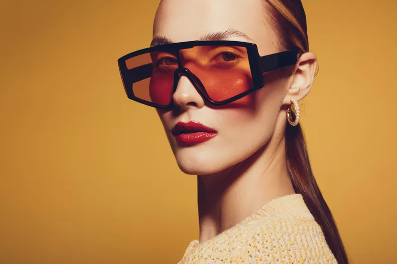 Stylish woman with slicked back hair wearing oversized orange sunglasses and gold hoop earrings poses against a warm yellow background. Fashion concept with bold accessories and makeup.