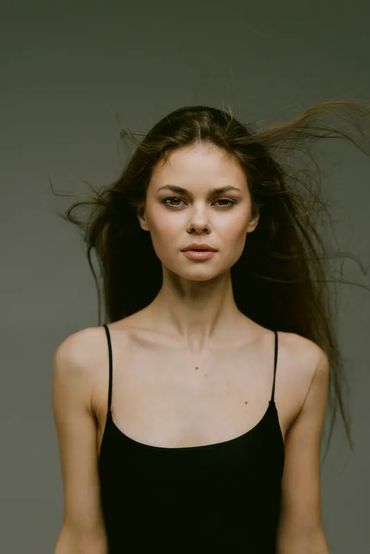 young woman with flowing hair wearing black tank top against gray background, calm and confident expression