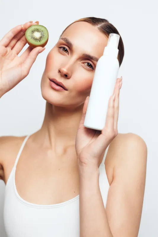 Young woman with clear skin holding a white cosmetic bottle and a sliced kiwi fruit. Concept of natural skincare, beauty routine, and healthy lifestyle on white background.