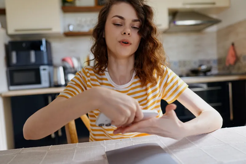 A woman in a striped shirt unboxes a package at home, standing by a sunny kitchen counter. Casual lifestyle moment showing anticipation and curiosity during home delivery.