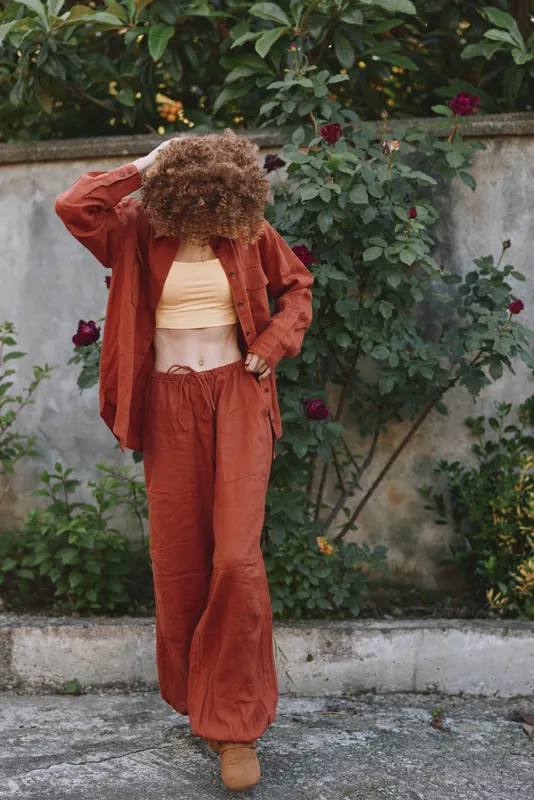 Woman lifestyle outdoors in rust orange outfit, casual pose with wideleg pants and cropped top, standing by greenery and wall, natural sunlight, relaxed mood with boho vibe