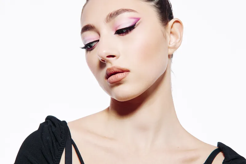 Young woman in creative fashion makeup poses with pink winged eyeliner and glossy lips styled for beauty photography on a clean white background.