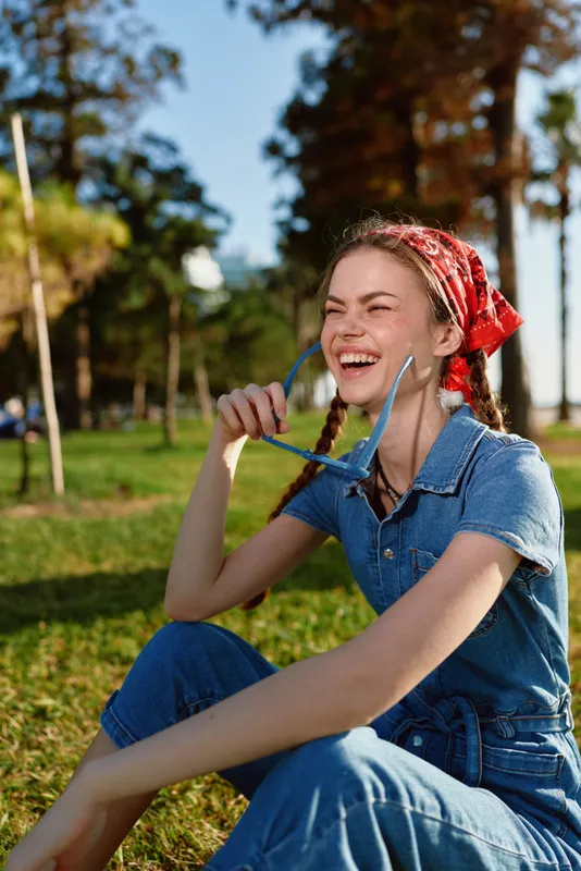 Lifestyle woman in modern boho-western denim street style laughing outdoors on sunny day, holding sunglasses with film color tone creating vibrant joyful mood.