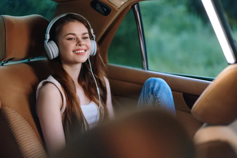 Young girl enjoying music in a car with headphones, smiling, showcasing joy and relaxation in a summer setting.