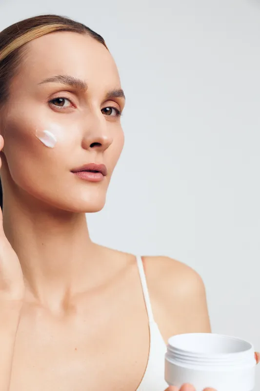 Young woman with short brunette hair applies moisturizing cream on her cheek in a bright studio with a calm and focused expression.