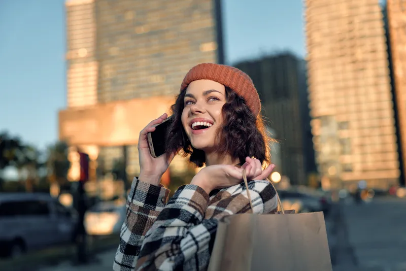 Woman shopping on city street, phone in hand and smile, beanie and bag in warm sunlight, portrait of urban joy with authenticity, candid lifestyle, golden hour glow, mindful living, emotional