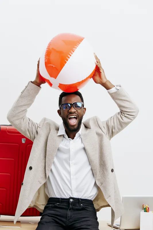 Joyful young man celebrating with a colorful beach ball indoors, casual business attire, smiling broadly with glasses, cheerful mood, modern office background, lifestyle concept.