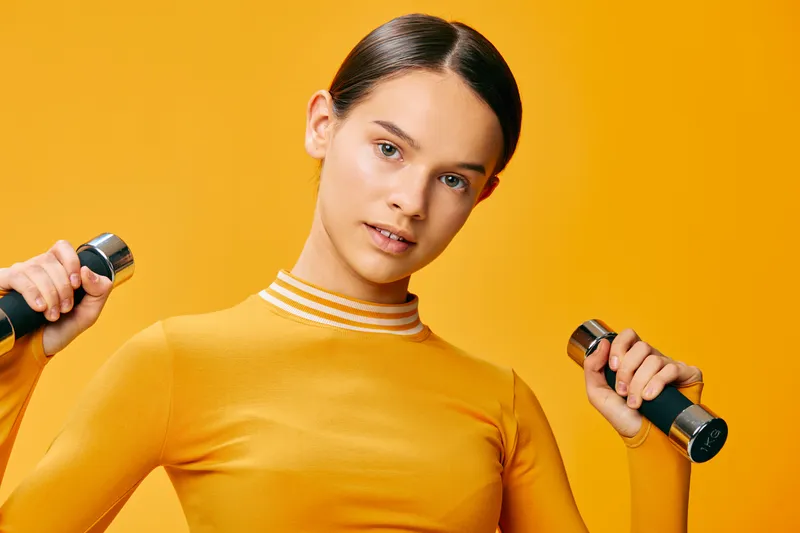 Woman fitness athlete lifting dumbbells against an orange studio backdrop, showcasing strength, balance, and focus during a high-energy workout session. Her yellow top complements the vibrant