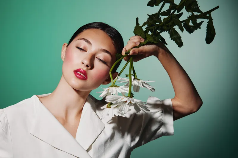 Attractive woman with glossy lipstick holds white daisies near her head, eyes closed, wearing an elegant white blouse. Studio shot on teal background. Concept of beauty, fashion, and creativity.