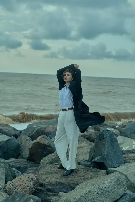 Woman standing on rocky shore, wind catching her coat, casual business attire, contemplating calm sea and open sky, outdoor style with movement, fashion and landscape elements, moody coastal scene