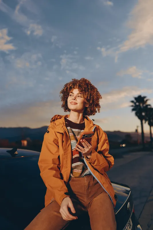 Woman with curly hair, orange jacket, sitting on scooter at sunset, outdoor lifestyle portrait, warm sky and palm silhouettes, relaxed travel mood