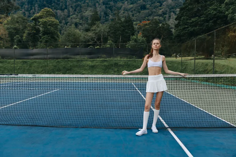 Young athletic woman in a sporty outfit poses confidently on a blue tennis court, showcasing summer energy and sporting spirit with green nature in the background