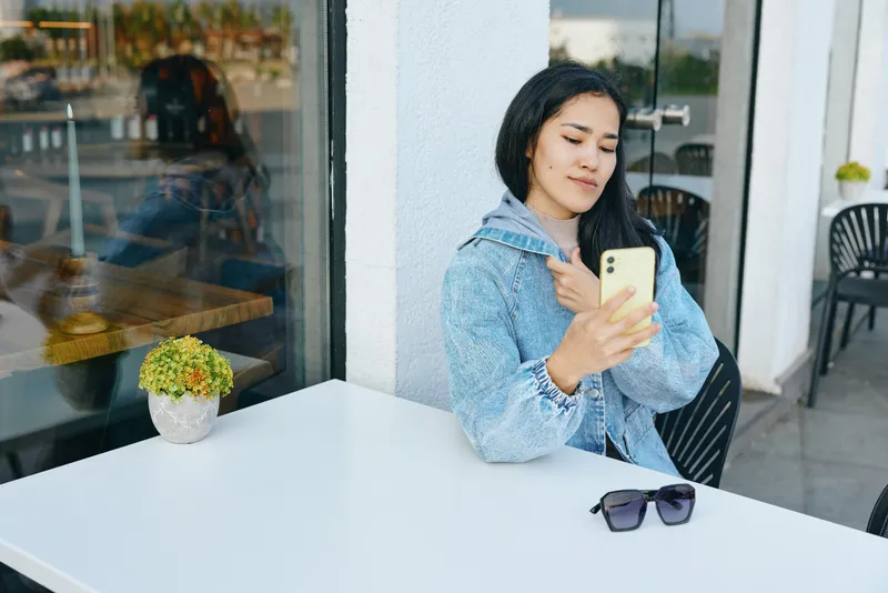 Woman in denim jacket sits at an outdoor cafe, glancing at her phone, beside a small plant and sunglasses on a white table, a calm urban moment captured in daylight.