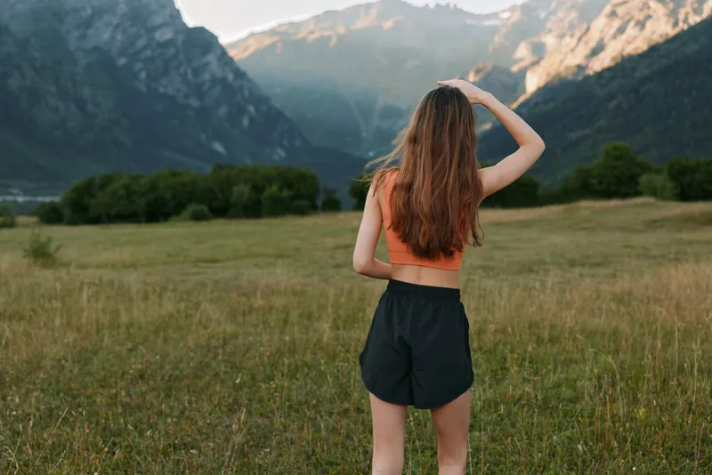 A young woman stands in a grassy field, gazing toward distant mountains under a clear sky. She wears a crop top and shorts, exuding a free, adventurous outdoor vibe.
