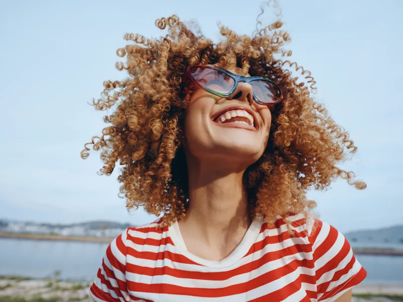 Joyful person with curly hair, wearing sunglasses and a red striped shirt, smiles brightly under a clear sky, with water and distant hills in the background.