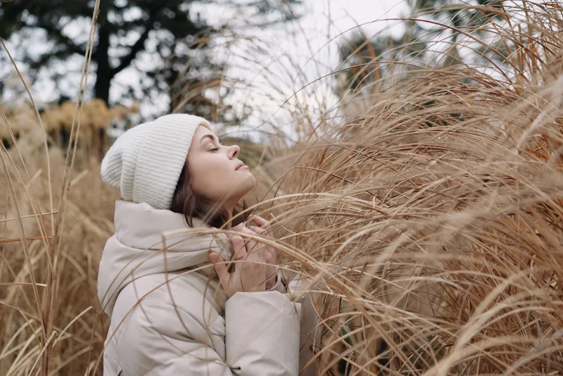 Woman in a warm white coat and beanie stands among tall dry grass, savoring the chilly breeze. A serene portrait highlighting winter textures, soft light, and a peaceful outdoor retreat.