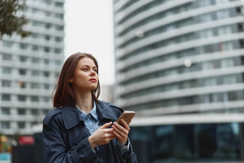 Young woman stands outdoors in a city setting, looking up with a smartphone in hand, casual jacket and denim shirt, modern street scene behind bright reflections behind softly.