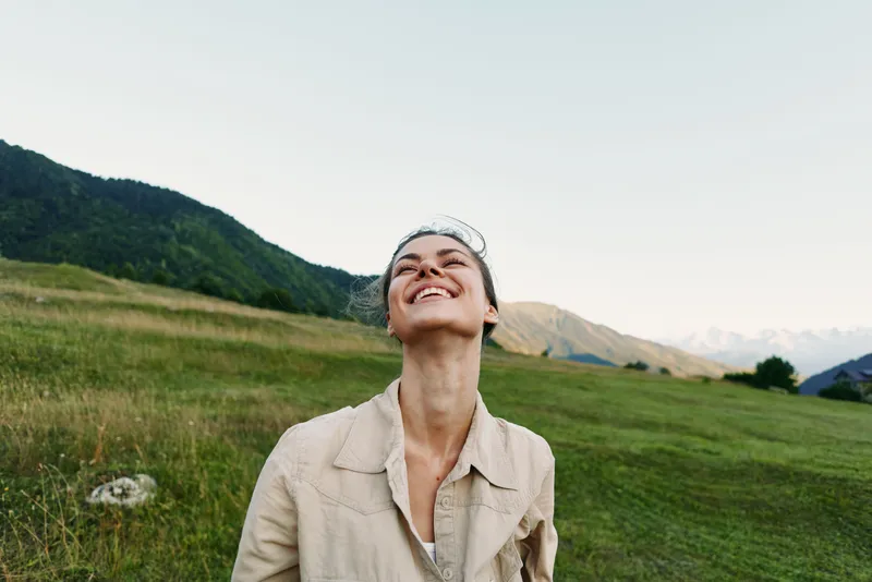 Young woman with short hair smiles joyfully while standing in a green meadow surrounded by mountains under a clear sky during daytime.