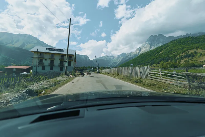 A scenic rural road winds through a mountainous valley, viewed from inside a car. Clear blue sky, rugged peaks, wooden fences, and small houses create a tranquil travel atmosphere.