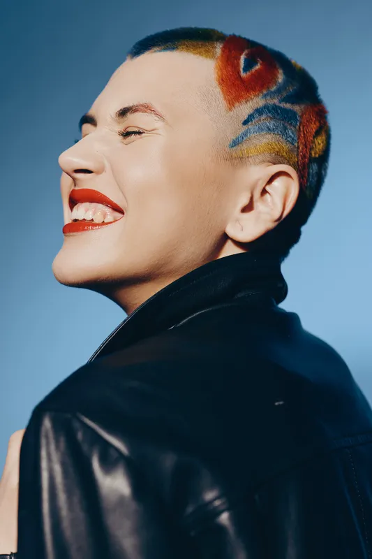 Smiling woman with colorful shaved hair and red lipstick wearing black leather jacket against blue background, expressing joy and individuality in a modern style.