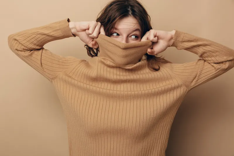 Young woman in beige knitted sweater pulls collar over her face, peeking with eyes. Neutral background and cozy clothing create a warm, playful mood and casual lifestyle concept.