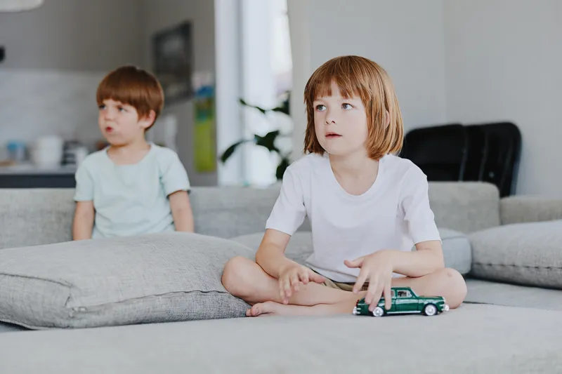 Focused girl playing with a toy car, sitting on a cozy sofa, surrounded by soft cushions and a modern interior, highlighting childhood joy and imagination.
