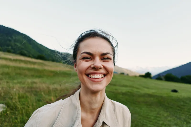 Young woman outdoors smiles broadly in a sunlit field with distant mountains, showcasing a relaxed, joyful lifestyle moment
