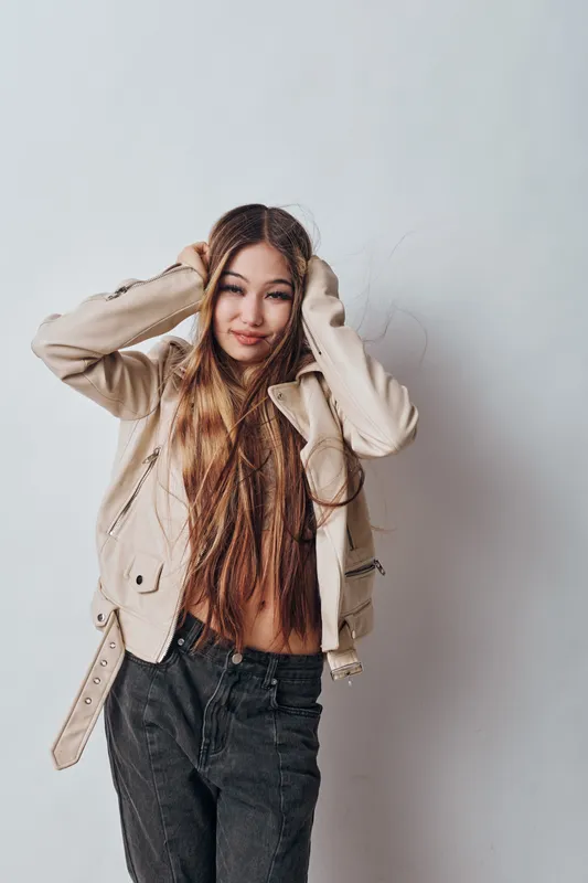 Young woman with long hair wearing beige jacket and black jeans posing with hands on head on light background. Casual outfit and relaxed expression in studio portrait.