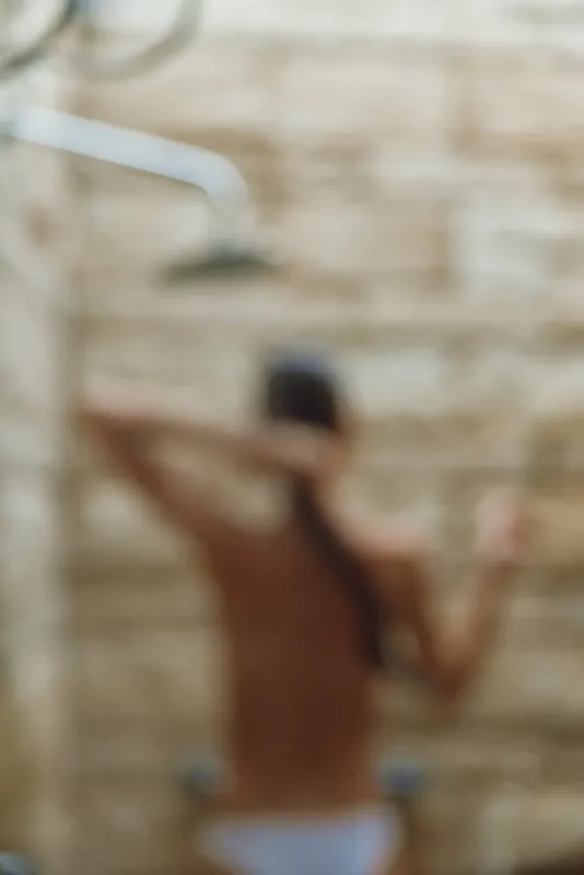 Woman showering outdoors under overhead shower with stone wall background, blurred artistic image capturing relaxation and natural bath setting.