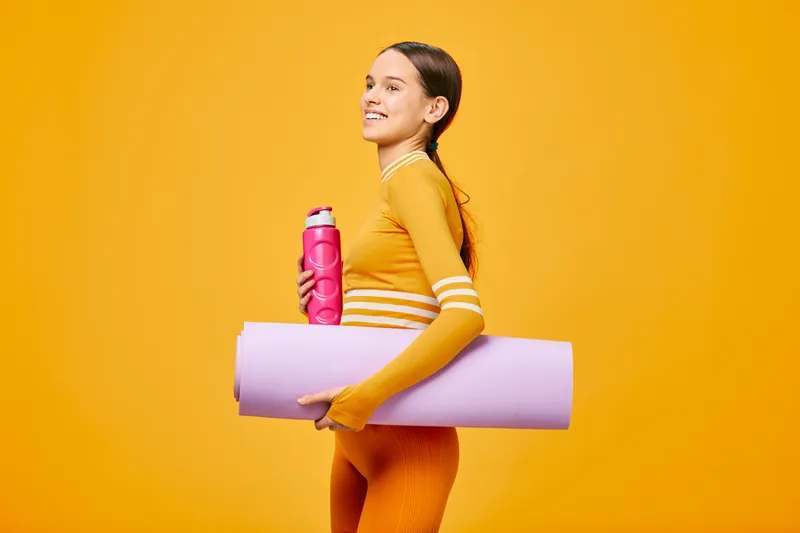 Woman in bright orange athletic wear carries a water bottle and a rolled yoga mat against a vibrant orange background, embodying fitness, sport and an active lifestyle.