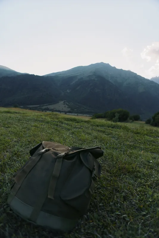 A lone backpack rests on a grassy meadow with distant mountains, inviting a sense of solitude and outdoor exploration under a clear sky.