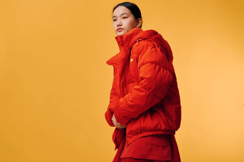 Woman in red puffer jacket standing against orange background with hands in pockets, looking thoughtfully to side with casual stylish winter outfit and confident posture.