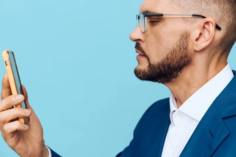 Professional man in a suit on an isolated colored background, gazing at a smartphone with calm focus and confident composure, conveying business readiness and thoughtful efficiency.