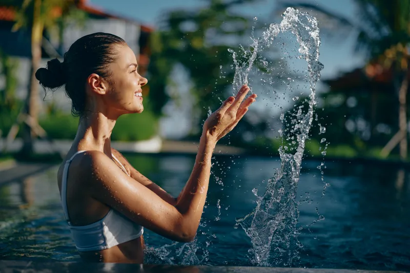 Smiling woman in a white top playfully reaches toward a water fountain in a pool, under sunny outdoors light, capturing a lifestyle moment of wellness, relaxation, joy, and refreshing mood.
