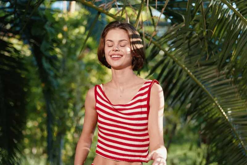 A cheerful young woman wearing a red and white striped crop top stands outdoors in a sunlit garden. She smiles with closed eyes, enjoying warm light and fresh air, conveying carefree happiness and