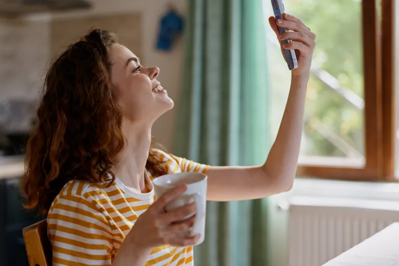 Smiling woman with curly hair holding a cup and raising a device in a bright home setting, a casual lifestyle portrait that emphasizes comfort, wellbeing, and everyday self care.