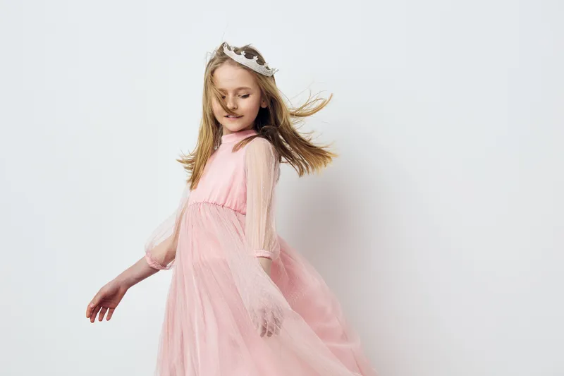A young girl in a pink tulle dress twirling joyfully, showcasing movement and elegance, with a playful expression and a crown in a light pastel background