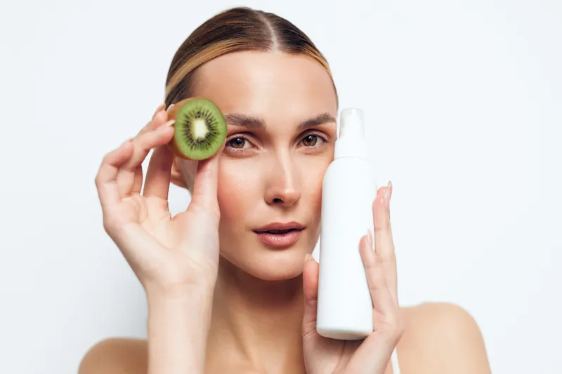 A young woman with clear skin holds a sliced kiwi near her face and a white skincare bottle in the other hand. Concept for natural beauty, skincare, and healthy lifestyle.