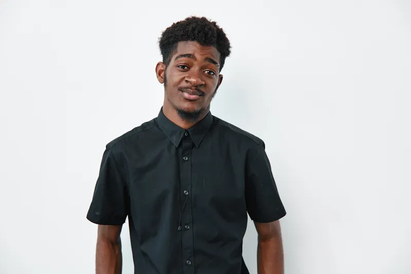 Young black man with dark skin wearing black short sleeve shirt standing against white background looking at camera with slight smile and relaxed posture.