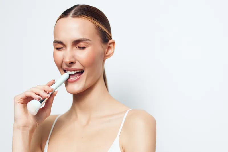 Young woman with closed eyes uses electric toothbrush for oral hygiene cleans teeth and gums with positive expression isolated on white background