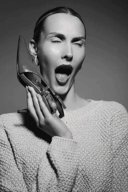 Young white woman with pale skin and slicked back hair holds high heel shoe near open mouth, eyes closed in dramatic expression. Fashion portrait in black and white studio lighting.