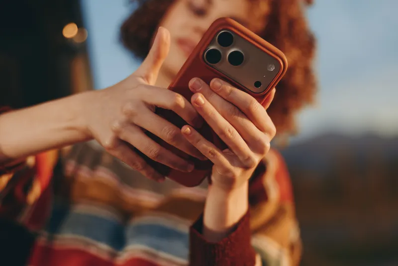 Woman with curly hair holds a smartphone in both hands outdoors. Close focus shows phone and fingers, with blurred background. Lifestyle communication moment, soft daylight, casual joy.