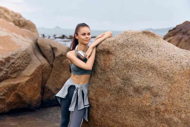 Fit young woman in sportswear enjoying a moment at the seaside, exuding confidence and relaxation against a rocky background, perfect for fitness and wellness themes