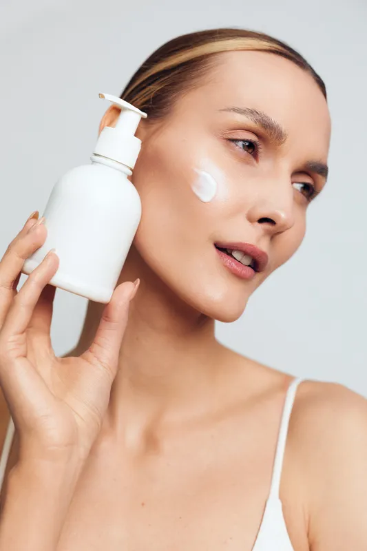 Young woman with clear skin holding white lotion bottle applying cream on cheek. Studio portrait with soft lighting on neutral background. Concept of skincare, beauty, moisturizing, wellness.