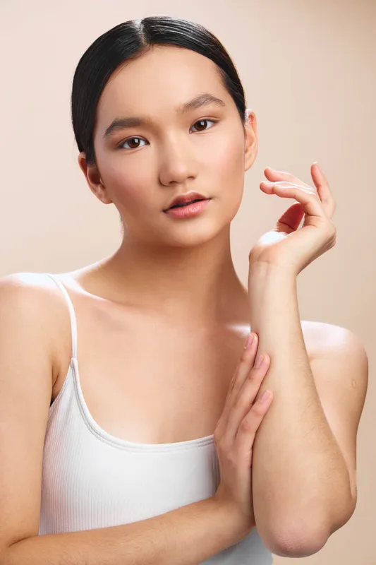 Asian woman with smooth skin rests her hand near her face in a simple studio portrait, calm expression and soft lighting evoke elegance, poise, and a timeless, professional aura.