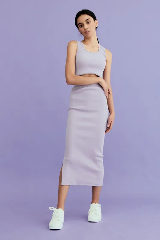young woman in lilac ribbed dress standing against purple background with casual sneakers, minimalistic style and relaxed confident pose, studio lighting